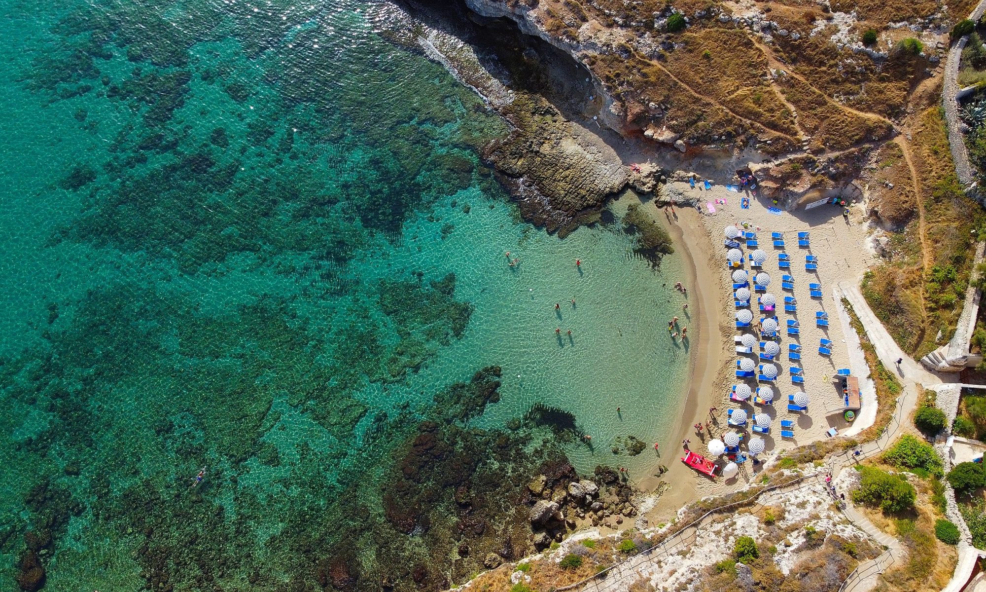 Spiaggia di Cala Corvino a Monopoli vista dall’alto, con sabbia dorata, ombrelloni blu e mare cristallino della Puglia.